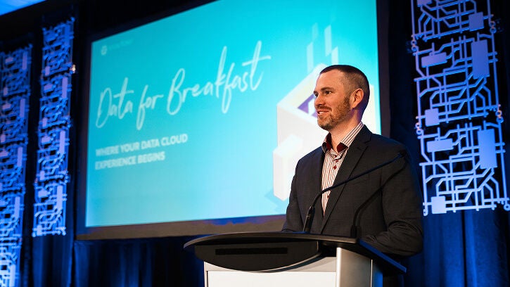 A smiling male speaker stands at a podium on a stage, ready to present at a Snowflake "Data for Breakfast" event. A large screen behind him displays the event logo.
