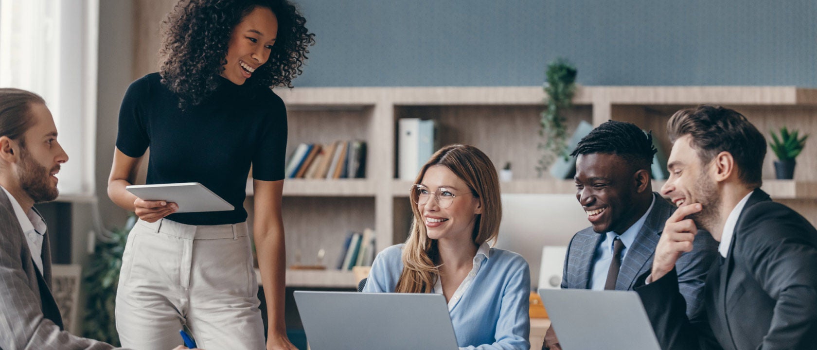 Five work colleagues discussing over two laptops. One woman has notebook in hand. 
