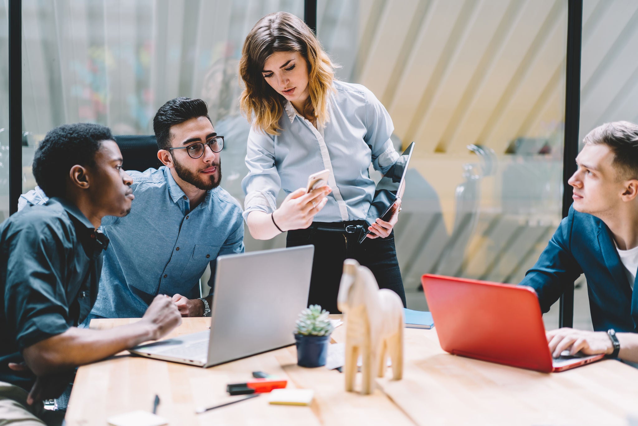 Young female with tablet showing data on smartphone to focused multiethnic male coworkers while working on project in meeting room of contemporary office