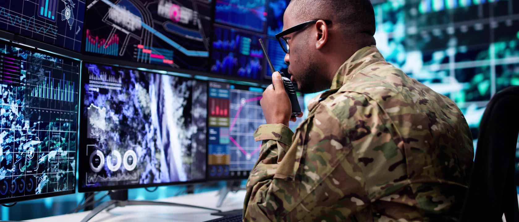 Soldier speaking into a walkie talkie while looking at three computer monitors