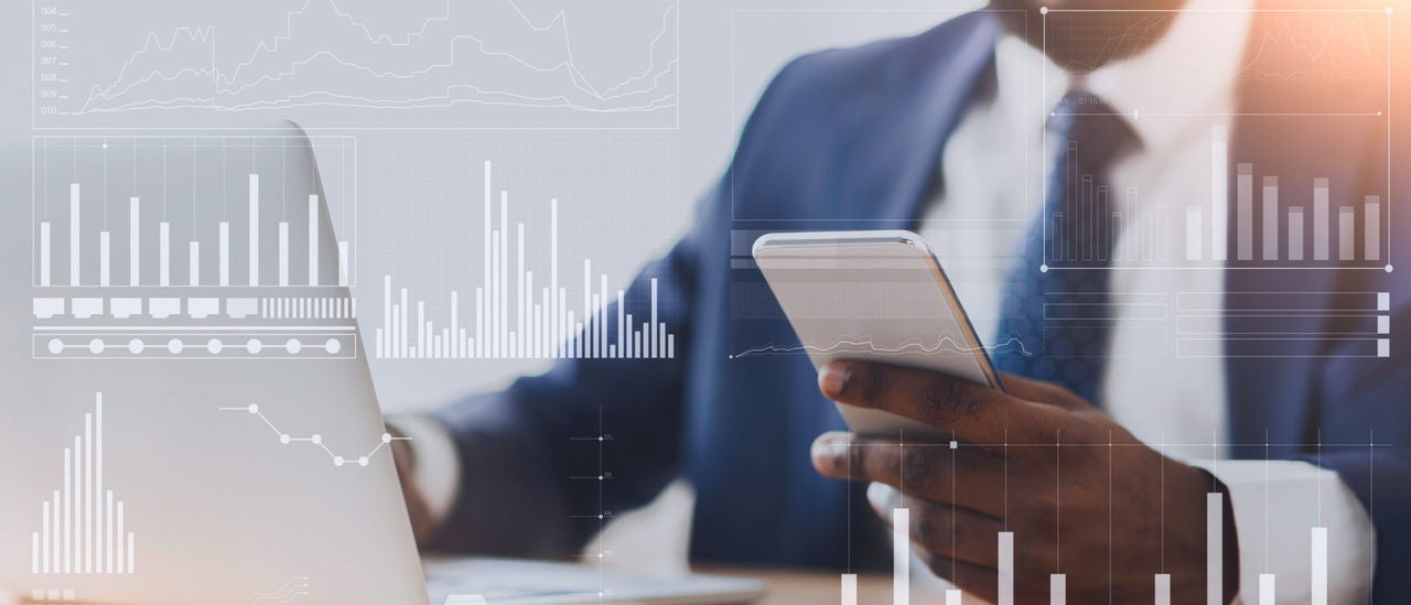 man wearing suit sitting at desk using phone in one hand and laptop; graph graphics overlay on image