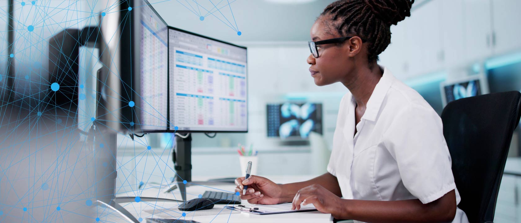 Photo of a woman in lab or medical clothing looking at multiple computer monitors full of metrics.