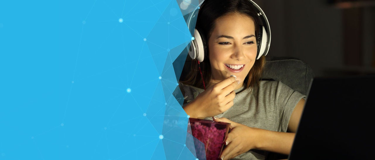 Woman wearing headphones, smiling, watching something while eating a bowl of popcorn