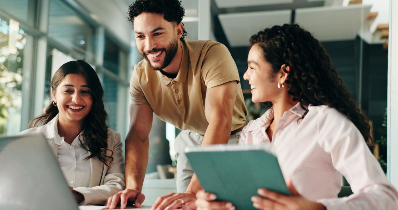Three colleagues, two women and a man, sit laughing at a table together while working