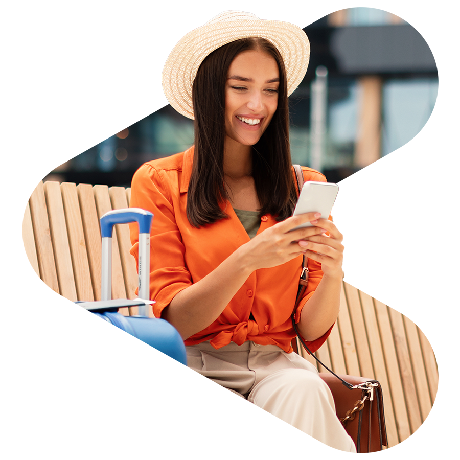 woman using phone sitting in airport next to her suit case; she is wearing a bright orange shirt and sun hat