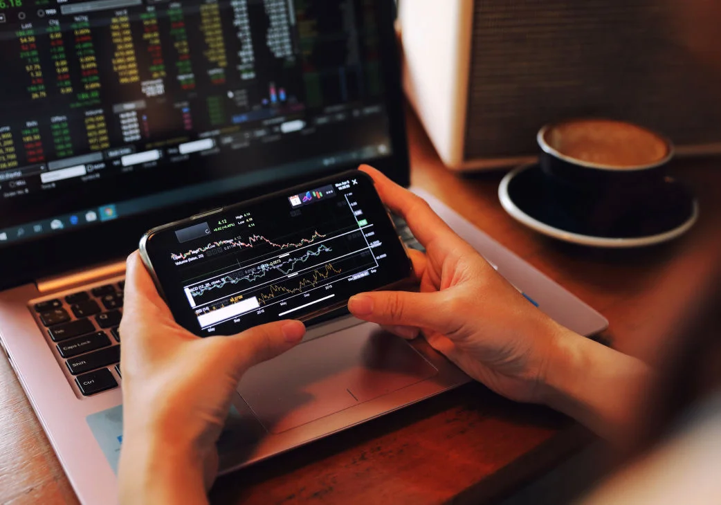 hands holding a phone in front of a laptop both showing financial charts