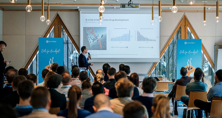 A presenter addresses an audience at a Snowflake "Data for Breakfast" event with a screen that displays related graphs and visuals.